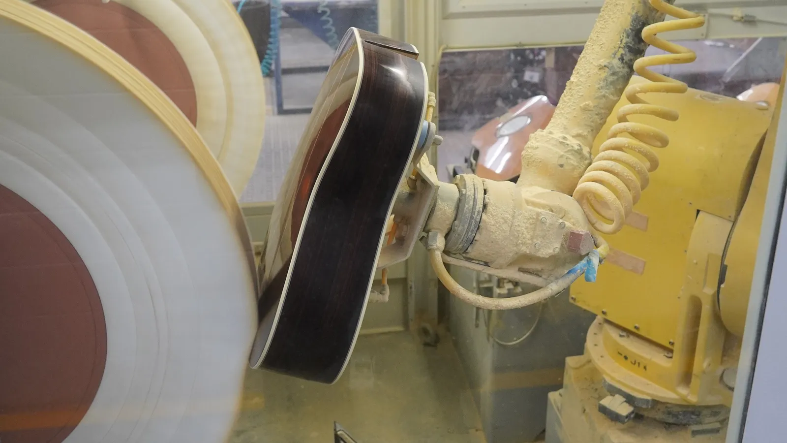 An industrial robot arm holding a guitar body against a sanding wheel in a factory