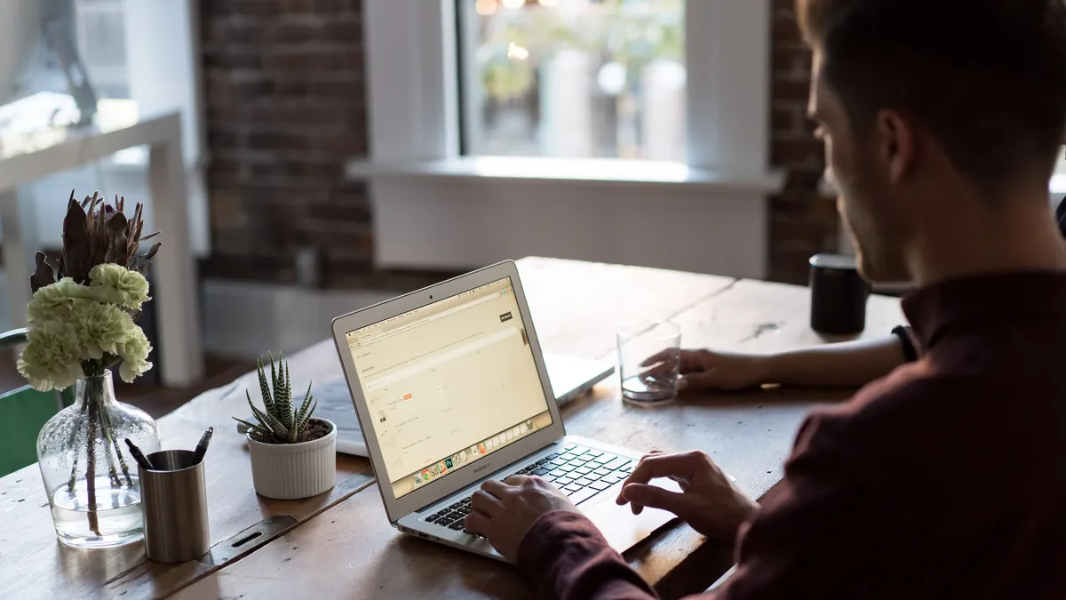 A man working on a laptop with a web browser open at a wooden desk in a bright office with a brick wall